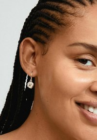 Boucle d'oreille en argent avec une pierre précieuse ronde et transparente, suspendue à un fil fin, portée par une personne aux cheveux tressés, vue en gros plan de l'oreille.