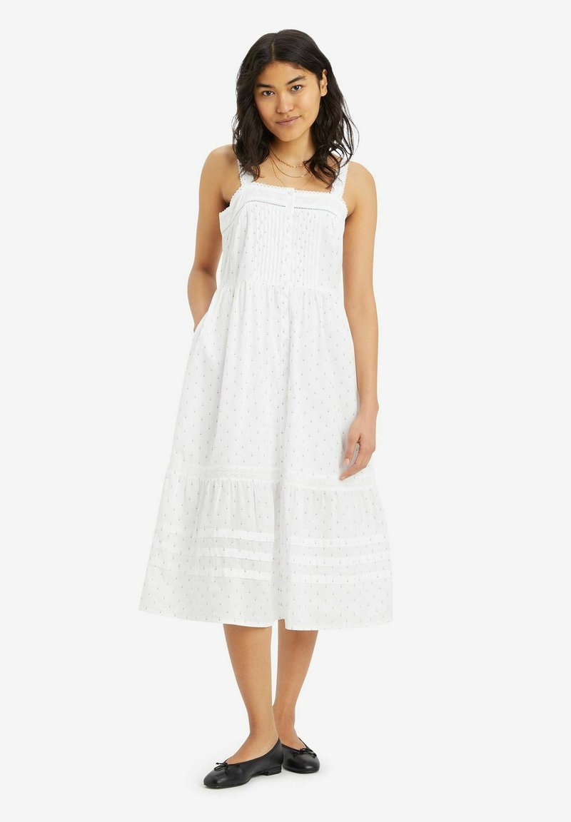 Weißes Midi-Kleid mit Spaghettiträgern, strukturiertem Stoff, Pünktchenmuster und gestuftem Saum. Tragen mit schwarzen Ballerinas.