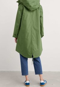 Grön kappa med huva, rundad fåll, insvängd midja och dragsko detaljer; bärs över blå cropped jeans och sneakers med blå accenter.