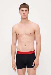 Schwarze Boxer Shorts mit einem roten Bund, der weißen Logo-Text trägt. Der Stoff wirkt glatt und passt eng an. Das Model ist oberkörperfrei.