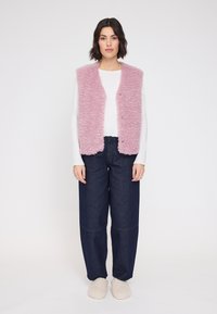 Faux-Fur-Weste in Pastellrosa, mit offenem Vorderteil, strukturiertem Oberflächen. Getragen über einem weißen Langarmshirt und kombiniert mit weit geschnittenen Jeans in dunklem Denim.