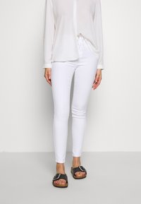 Jeans blancos ajustados con un acabado de tela suave, combinados con una blusa blanca de manga larga y sandalias negras con una gran hebilla.