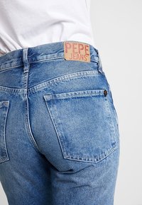 Ljusblå denimjeans med en strukturerad yta, med en synlig bakficka och ett märkeslapp i läder med "PEPE JEANS" på midjebandet.
