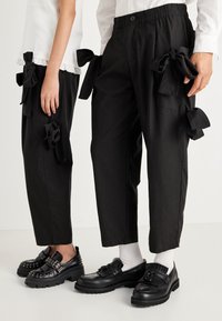 Calças pretas curtas com bolsos laterais adornados com grandes laços de tecido. O tecido parece suave; cós elástico com um botão. O calçado inclui sapatos pretos.