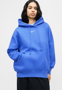 Νεαρό άτομο που φοράει φωτεινή μπλε μπλούζα Nike με κουκούλα και έχει τα χέρια του στην τσέπη μπροστά, μπροστά σε ένα απλό φόντο.