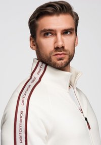 Chaqueta blanca con cremallera, con acentos de rayas rojas y marrones y texto en las mangas, que presenta un cuello alto y una textura suave.