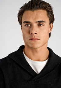 Schwarzer gestrickter Pullover mit Schalkragen, getragen über einem weißen T-Shirt. Verfügt über einen strukturierten Stoff und eine neutrale Farbpalette.