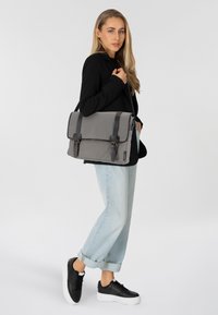 Bolso mensajero gris con correas de cuero negro, cierre con solapa y hebillas. Presentado con una mujer vestida con una chaqueta negra y vaqueros azules.