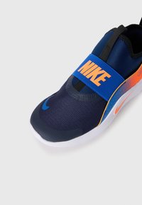 Chaussure de sport marine avec le logo "NIKE" orange, une sangle extensible, une tige en mesh texturé et une semelle blanche. Présente un dégradé de bleu à orange près du talon.