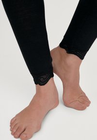 Leggings neri a coste con un bordo di pizzo alla caviglia, caratterizzati da un tessuto testurizzato e un design aderente, appoggiati sopra piedi nudi.