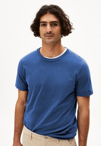 Blauwe katoenen T-shirt met korte mouwen en een ronde halslijn, getoond op een model dat beige broek draagt. Glad textuur, casual pasvorm.