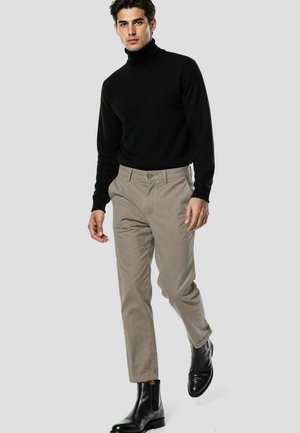 Mann geht, trägt schwarzen Rollkragenpullover, beige Hose und schwarze Leder-Stiefeletten vor einfachem hellem Hintergrund.