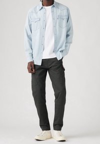 Helles blaues Denimhemd mit Druckknöpfen, darunter ein weißes T-Shirt mit Rundhalsausschnitt, dunkle Cargo-Hosen und beigefarbene Sneakers. Zwei sichtbare Brusttaschen.