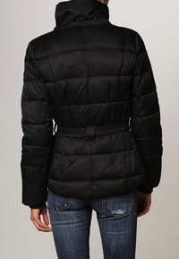 Sort pufferjakke med høj krave, quiltet design og justerbart bælte i taljen. Båret med blå jeans, viser bagsiden.