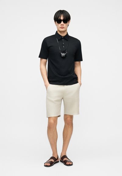 Άνδρας που φοράει μαύρο πόλο πουκάμισο, μπεζ σορτς, σκουρόχρωμα γυαλιά ηλίου, μαύρο κολιέ με μικρή θήκη και καφέ σανδάλια, στέκεται με τα χέρια στις τσέπες.