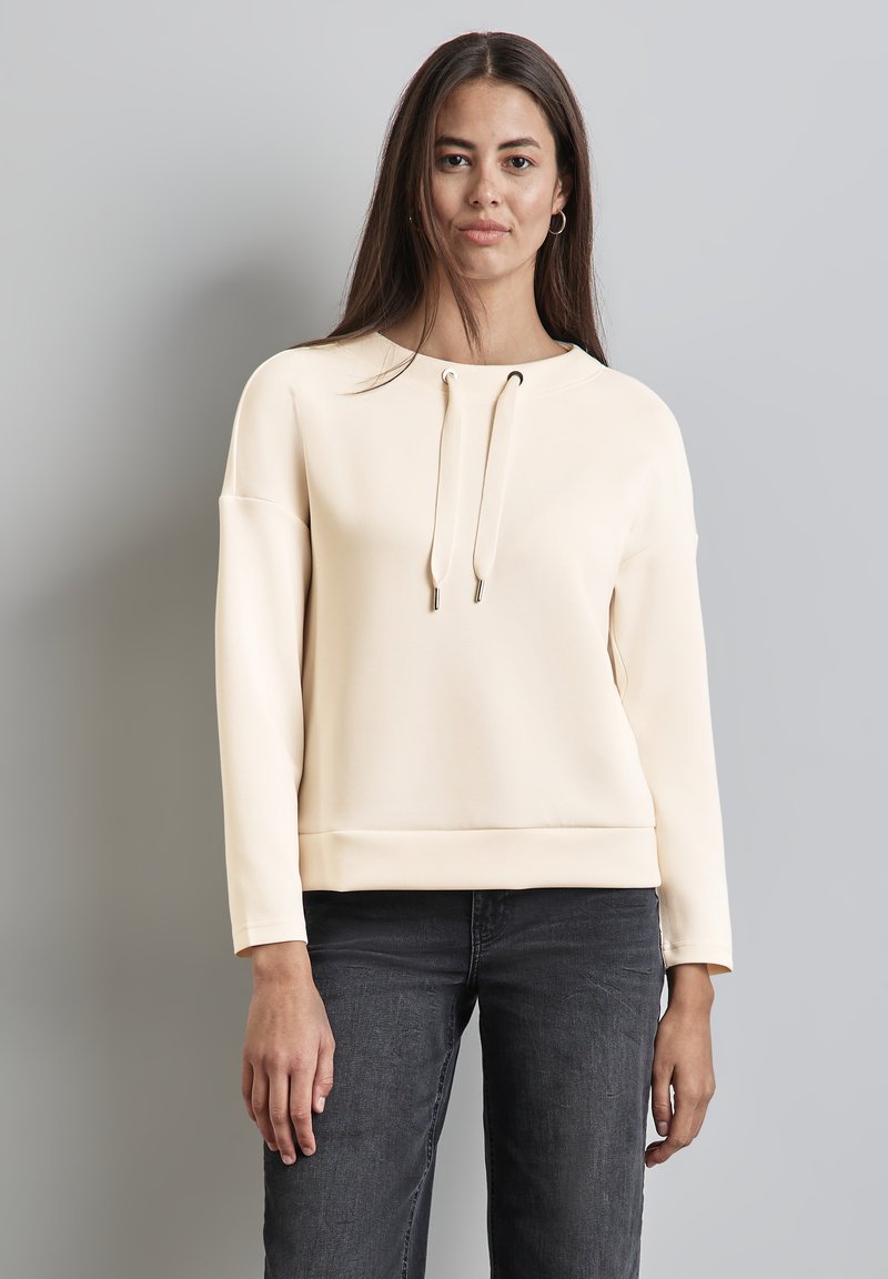 Felpa beige chiaro con scollatura rotonda, chiusura con coulisse e maniche lunghe. Tessuto morbido con vestibilità rilassata e orlo a costine.