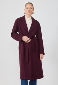 Cappotto in lana bordeaux con cintura, ampie alette e maniche lunghe, abbinato a una maglietta bianca e jeans blu. Tessuto morbido, design lungo fino al ginocchio.