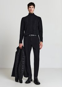 Schwarzer, gerippter Rollkragenpullover, maßgeschneiderte schwarze Hose und eine schwarze Lederjacke. Schwarze Stiefel vervollständigen das Outfit und betonen den monochromen Look.