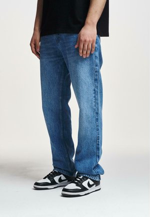 Light blue denim jeans with a relaxed fit, visible pockets, and slight fading. Paired with black and white sneakers.