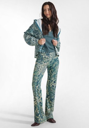 Kvinde iført teal-mønstret jakke og matchende udvidede bukser over en teal top, stående og justerende jakkraven, hvid baggrund.