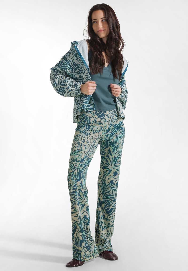 Frau trägt eine türkis gemusterte Jacke und passende ausgestellte Hose über einem türkisfarbenen Oberteil, steht und richtet den Jackenkragen, weißer Hintergrund.