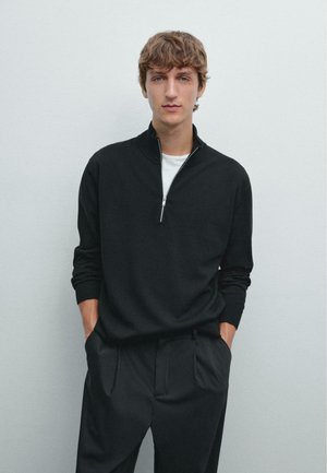 Maglione nero con zip e colletto alto, realizzato in un tessuto morbido. Modello abbinato a pantaloni neri, mani in tasca, in piedi davanti a uno sfondo semplice.