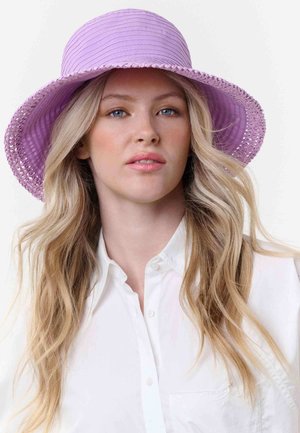 Blonde Frau mit welligem Haar trägt einen breitkrempigen lavendelfarbenen Hut und ein weißes Hemd mit Knöpfen und blickt mit neutralem Ausdruck nach vorne.