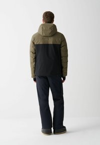 Padded jacket s kapucňou, olivovozelená horná časť, čierna dolná časť. Čierne nohavice, robustné topánky so svetlohnedou podrážkou.