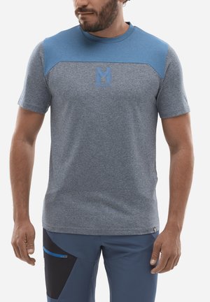 Homme portant une chemise à manches courtes bleu-gris avec un logo sur la poitrine et un pantalon bleu avec une poche noire et un détail de fermeture éclair bleu.