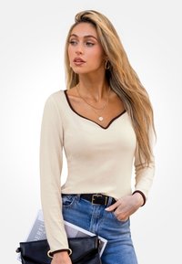 Top a maniche lunghe a coste di colore avorio con scollatura a V bordata in marrone, abbinato a jeans blu a vita alta e una pochette nera.