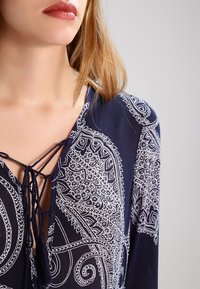 Námořnická modrá blůza s vzorem, která má výstřih do V se šněrovacím detailem a bílým paisley designem na jedné straně. Měkká tkanina s hladkou texturou.