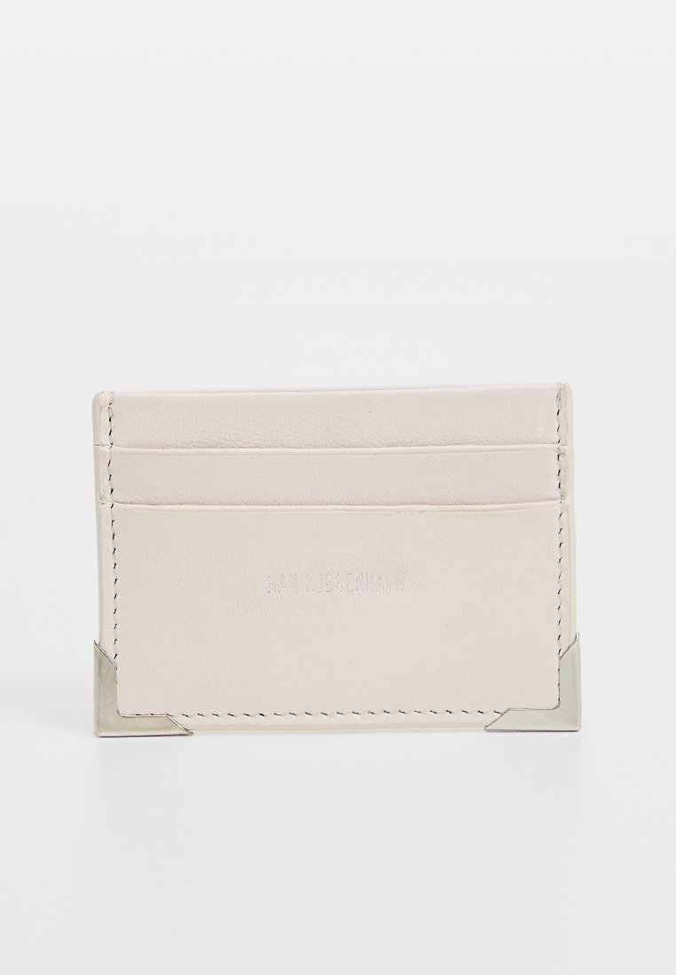 Porte-carte en cuir beige avec quatre fentes pour cartes, bords cousus et accents métalliques argentés dans les coins inférieurs. Logo de la marque en relief sur le devant.