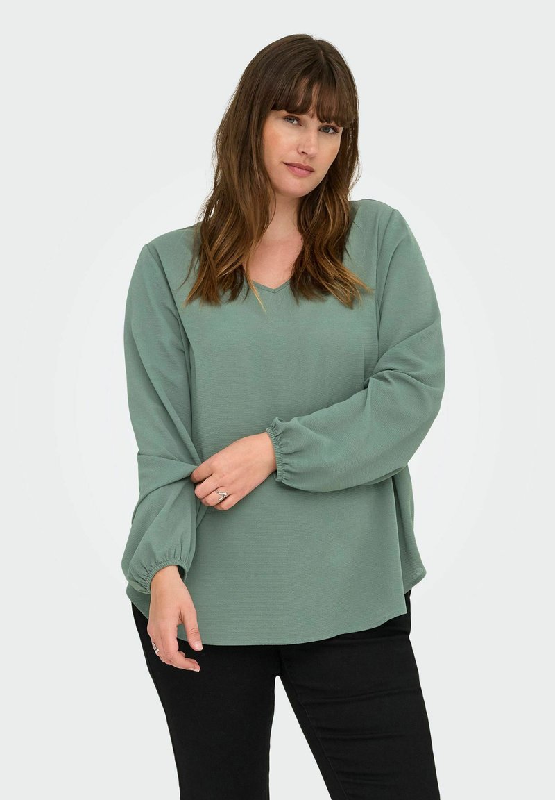 Minta zaļas krāsas long-sleeve tops ar V-veida kaklu, atvieglu piegriezumu un elastīgām piedurknēm, izgatavots no maigām, teksturētām audumam. Valkāts kopā ar melnām biksēm.