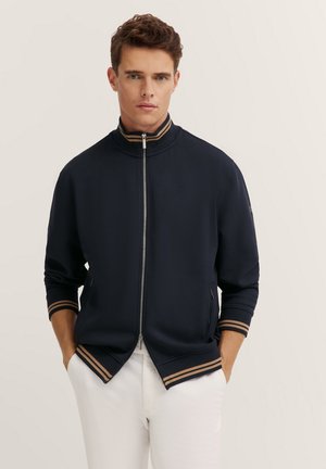 Navy zip-up jacket with a high collar, featuring gold and brown striped accents on cuffs and hem, made of smooth fabric. Side pockets included.