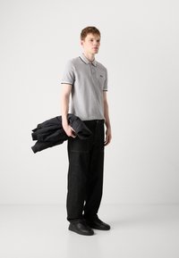 Γκρι polo μπλουζάκι με αντίθεση μαύρες λεπτομέρειες, σκούρες φαρδιές τζιν παντελόνες και μαύρα αθλητικά παπούτσια. Κρατάει μια μαύρη ζακέτα. Απλός, σύγχρονος σχεδιασμός.