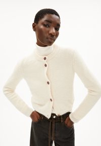 Cardigan in maglia color crema con colletto arrotondato, cinque bottoni marroni e bordi a costine, abbinato a jeans scuri. Texture morbida.