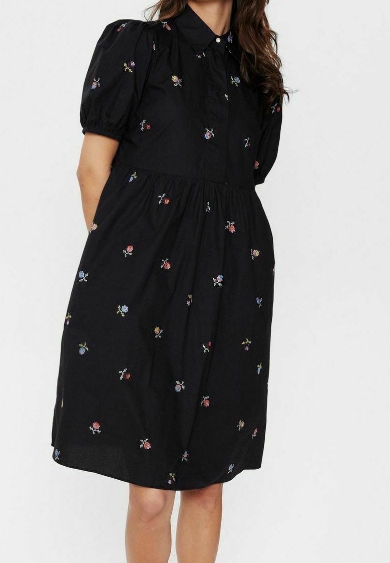 Femme portant une robe noire mi-longue avec des manches courtes bouffantes et une petite broderie florale multicolore.