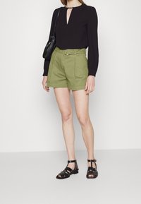 Blusa nera a maniche lunghe con scollatura a chiave, pantaloni corti a vita alta verde oliva con passanti per cintura e sandali neri con cinturini.