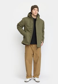 Olive green insulated jacket with a high collar and zipper, paired with beige cargo pants and white sneakers. Brown knit beanie on head.