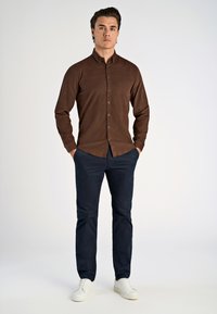 Bruine lange mouwen overhemd met een subtiele textuur, gecombineerd met marineblauwe chinos en witte sneakers. Het model staat met handen in de zakken.