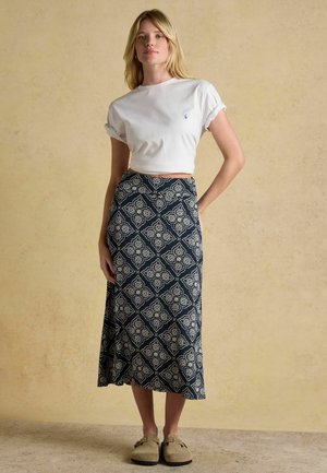Young woman standing with one hand on hip wearing a white cropped t-shirt, patterned navy midi skirt, and beige slip-on shoes.