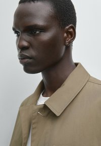 Tan jacket met een kraag, gemaakt van een gladde stof. Gedragen over een wit shirt, zijwaarts getoond, waarbij de naden en textuur van de jas worden benadrukt.