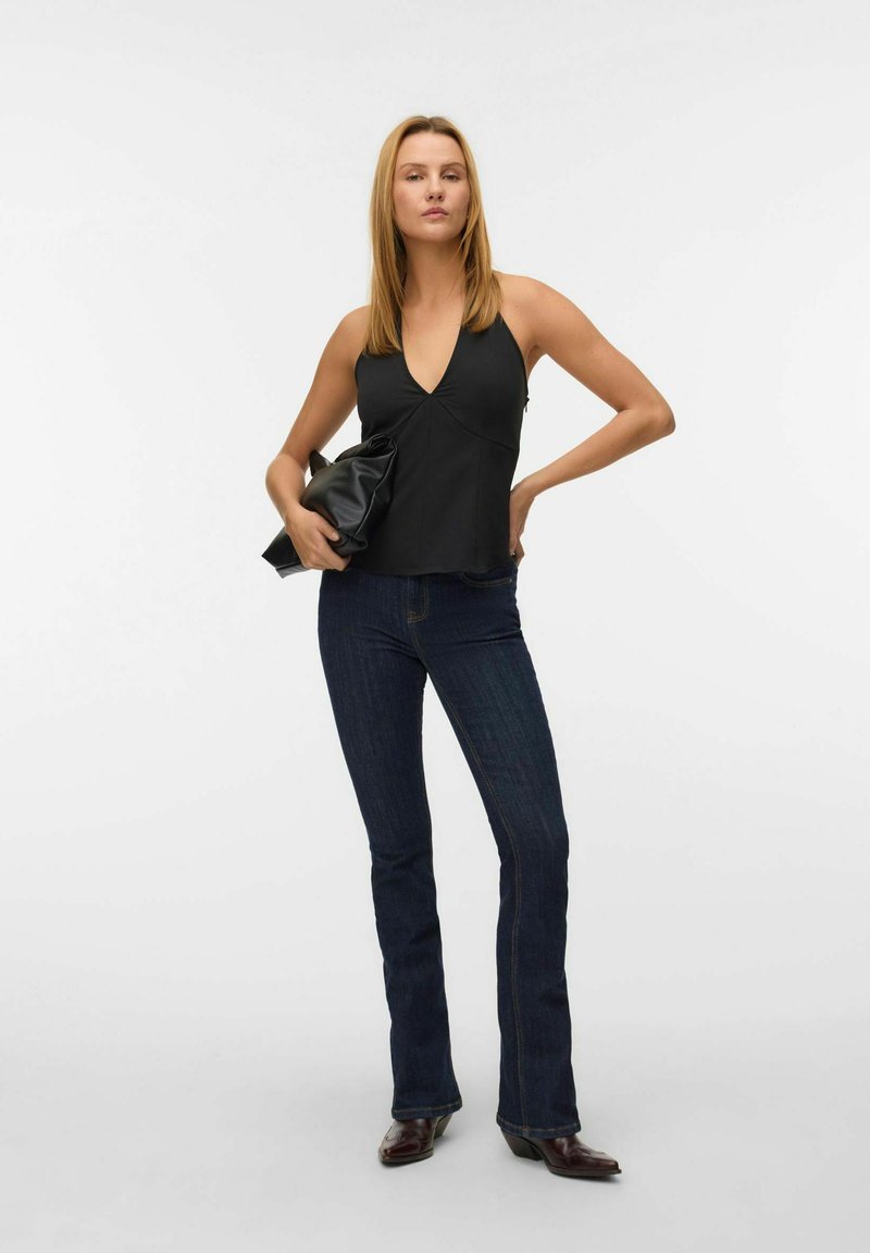 Zwarte haltertop met een diep decolleté, gecombineerd met donkere broek met een flared model en glanzende bruine enkellaarsjes. Model houdt een zwarte clutch vast.