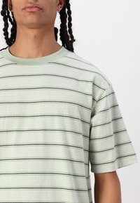 Camiseta verde a rayas con rayas horizontales de diferentes anchos. Material suave con cuello redondo y mangas cortas.