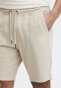 Pantalones cortos de algodón beige con cinturilla ajustable, bolsillos laterales y un logotipo gráfico de color claro en la pierna izquierda. Textura suave.