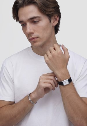Mladý muž s hnědými vlasy si na levé zápěstí upravuje černý kožený hodinkový pásek, má na sobě bílé tričko a na pravém zápěstí stříbrný náramek, za ním neutrální pozadí.