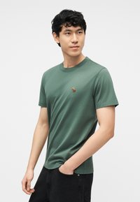 Νεαρός άνδρας με κοντά μαύρα μαλλιά που φοράει πράσινο μπλουζάκι με στρογγυλή λαιμόκοψη και ένα μικρό κεντητό ζωάκι, με το χέρι στην τσέπη μαύρου παντελονιού.