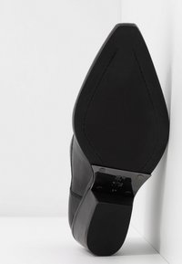 Chaussure en cuir noir avec un bout pointu et une tige lisse. Présente un talon structuré et une semelle texturée noire. Accents métalliques visibles.