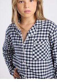 Chemise de pyjama en flanelle à carreaux bleu marine et blanc avec une poche poitrine, une fermeture boutonnée sur le devant, et un passepoil contrastant le long du col et de la patte de boutonnage.