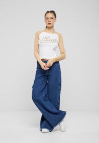 Hvit crop-top med brune ermer som har "Kani" grafikk, kombinert med vide blå denim cargo-bukser med frynset kant og sidelommer.